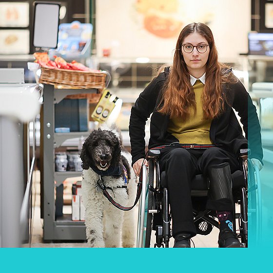 ein Hund begleitet eine Rollstuhlfahrerin beim Einkaufen
